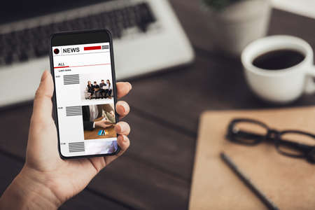 Female hand holding smartphone with last news articles on screen at table copy space collage
