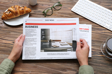 Woman reading newspaper at workplace with croissant on wooden background